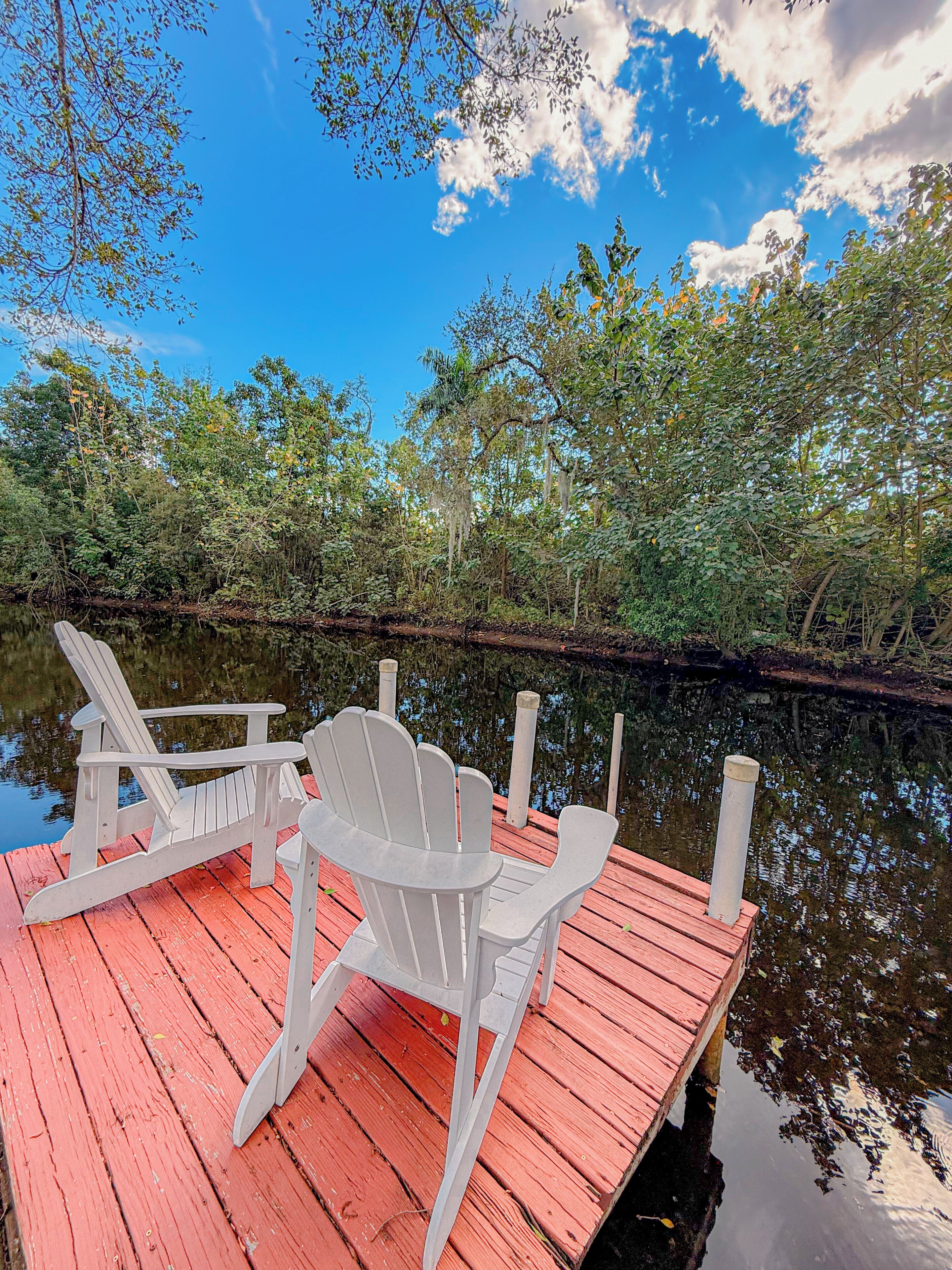 Une jetée en bois avec deux chaises Adirondack blanches, donnant sur une étendue d’eau calme, le tout entouré d’une végétation luxuriante et d’arbres.