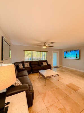 A living room with a sectional sofa, a coffee table, a ceiling fan, a TV, and a large window with blinds. - North Breeze, lake view with heated Pool (North Fort Myers)