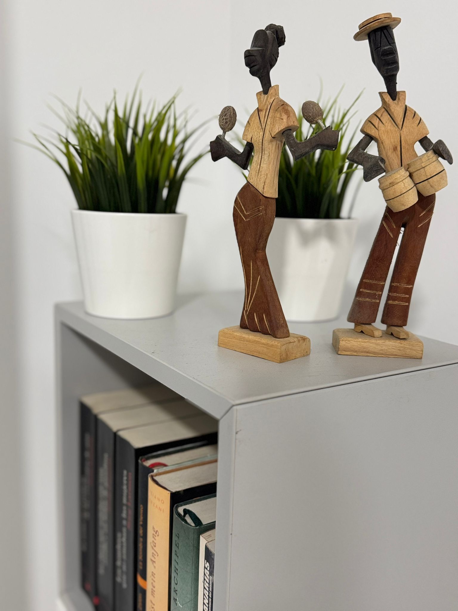 Due sculture in legno su uno scaffale con libri e piante in vaso.