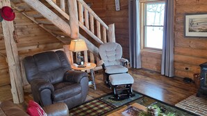 A cozy cabin interior with a wooden staircase, leather furniture, a lamp, and a window with curtains.
