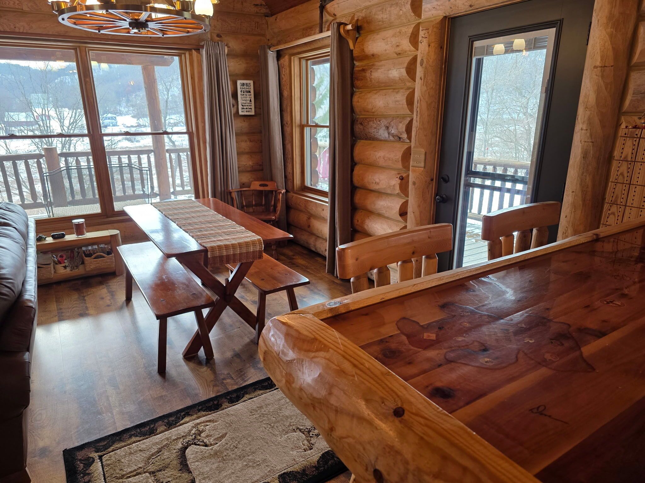 A log cabin interior with a wooden dining table, a sofa, and a view of a snowy landscape through large windows.