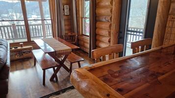 A log cabin interior with a wooden dining table, a sofa, and a view of a snowy landscape through large windows.