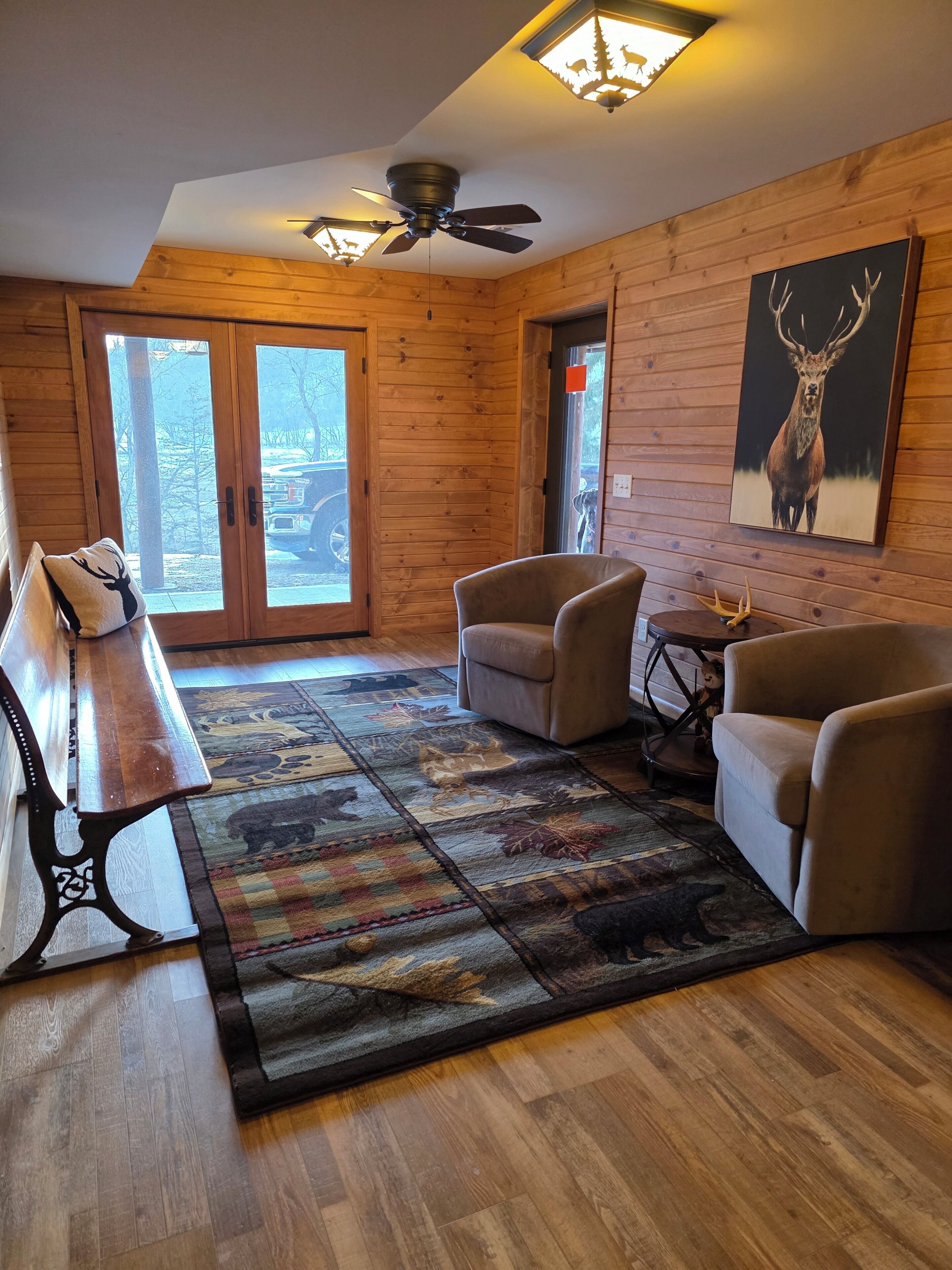A wooden interior with a ceiling fan, a bench, two armchairs, a rug, and a deer painting.