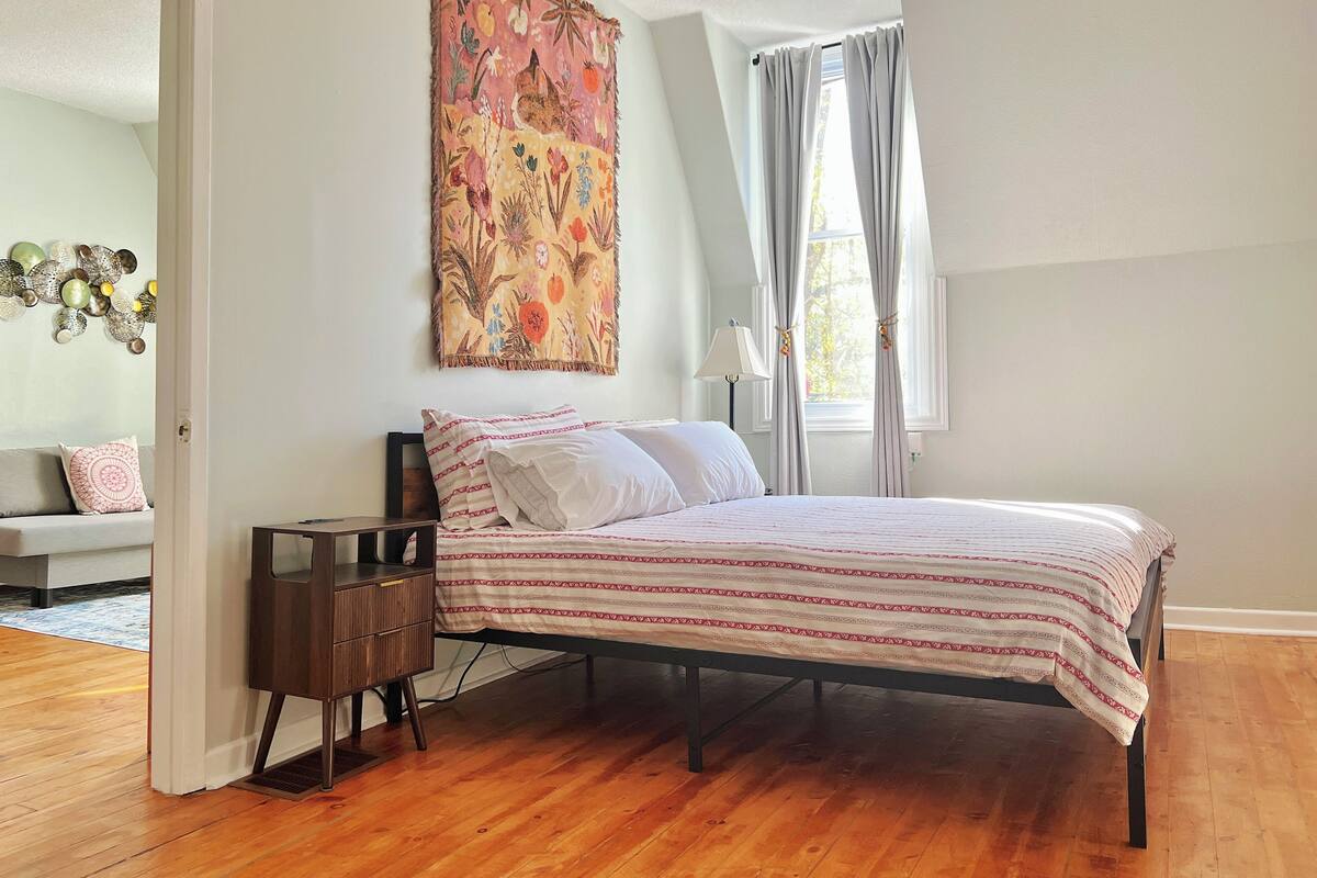 Un dormitorio con cama, mesita de noche, tapiz en la pared, ventana con cortinas y puerta que da a otra habitación.