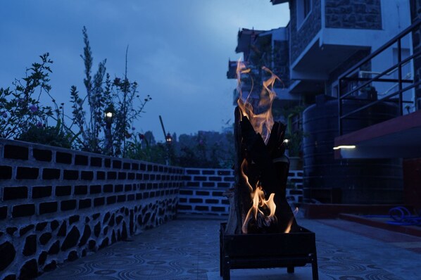 A fire burning in a metal container on a patio with a brick wall and a building in the background.