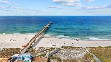 A beach with a long pier extending into the ocean, a sandy beach area with umbrellas, and a small building with a blue roof.