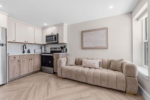 A modern kitchen with white cabinets, a microwave, and a stove. There is a beige sofa with cushions and a framed abstract artwork on the wall. - Modern Queen Studio | Steps to the Beach (Panama City Beach)