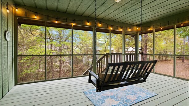 A covered porch with a swing, wooden flooring, and a view of trees. - Salty Clover𓇼Vintage Vibes + Modern Comfort (Oak Island)