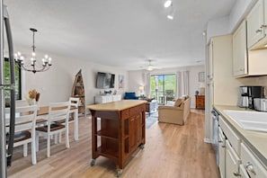 A spacious kitchen with a central island, a dining area with white chairs, a sofa, and a TV mounted on the wall. - Salty Clover𓇼Vintage Vibes + Modern Comfort (Oak Island)