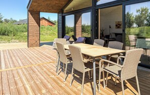 Eine Holzdielen-Terrasse mit Tisch und Stühlen, eine Glastür zum Innenbereich und Blick auf ein Holzgebäude sowie Grünflächen.