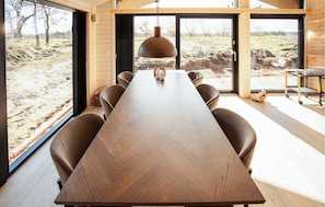 A long wooden table with chairs, a hanging pendant light, and large windows with a view of an outdoor landscape.