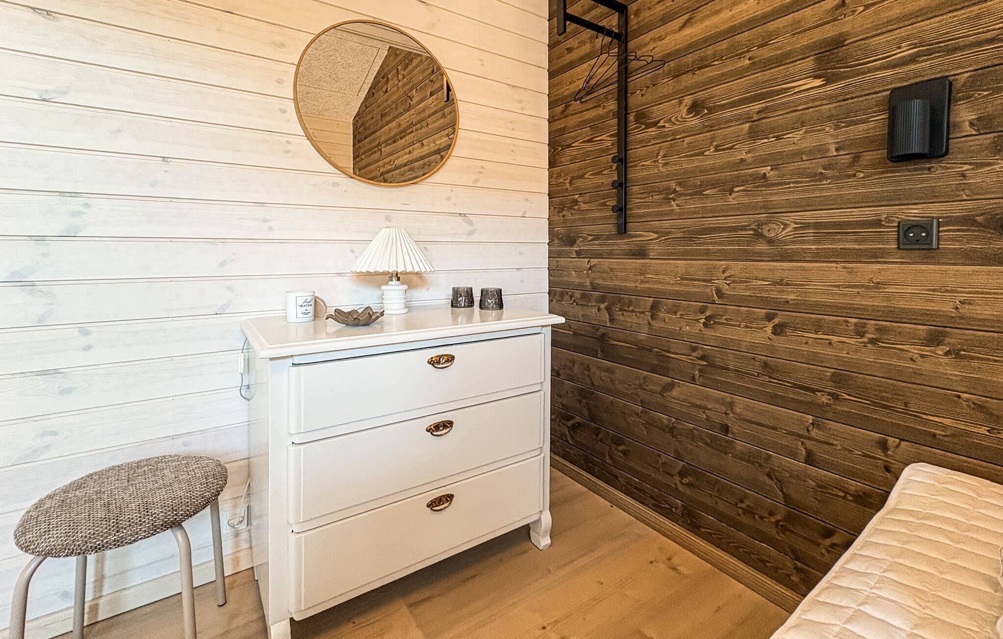 A room with a white dresser, a round mirror, a small stool, and a wooden wall.