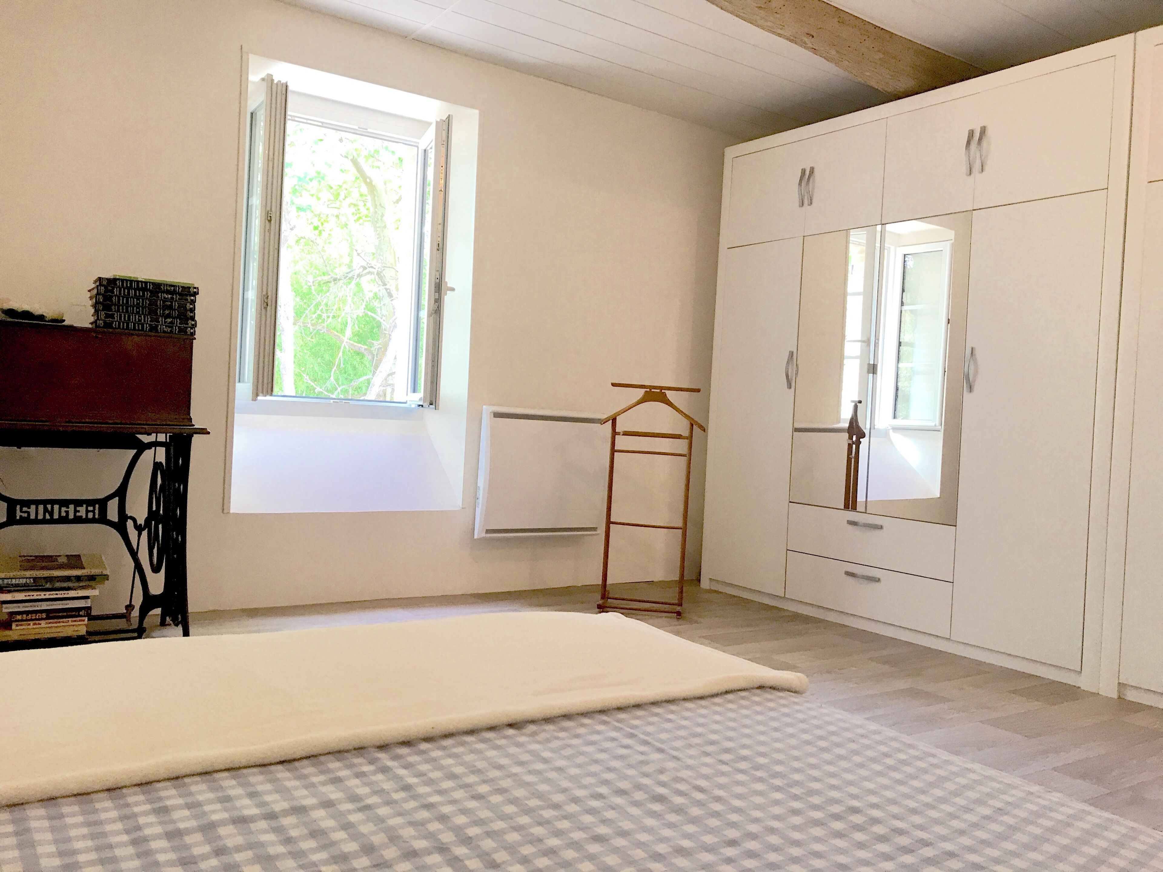A room with a large wardrobe, a radiator, a window with a view of greenery, and a bed with a checkered bedspread.