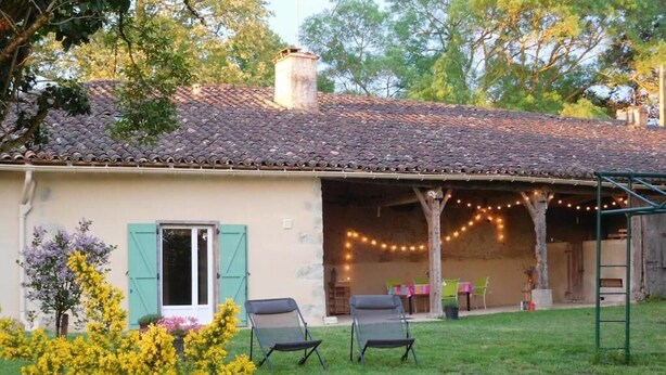 A house with a tiled roof, a covered patio with string lights, and a grassy yard with two chairs.