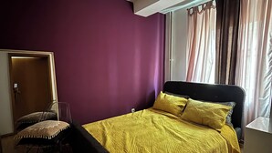 A bedroom with a bed, a chair, a desk, a window with curtains, and a ceiling light. - Cozy apartment in a vibrant neighborhood  with fast internet (Skopje)