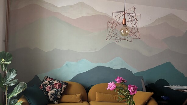 A modern living room with a sofa, a coffee table with papers, a vase with flowers, and a wall mural. - Cozy apartment in a vibrant neighborhood  with fast internet (Skopje)