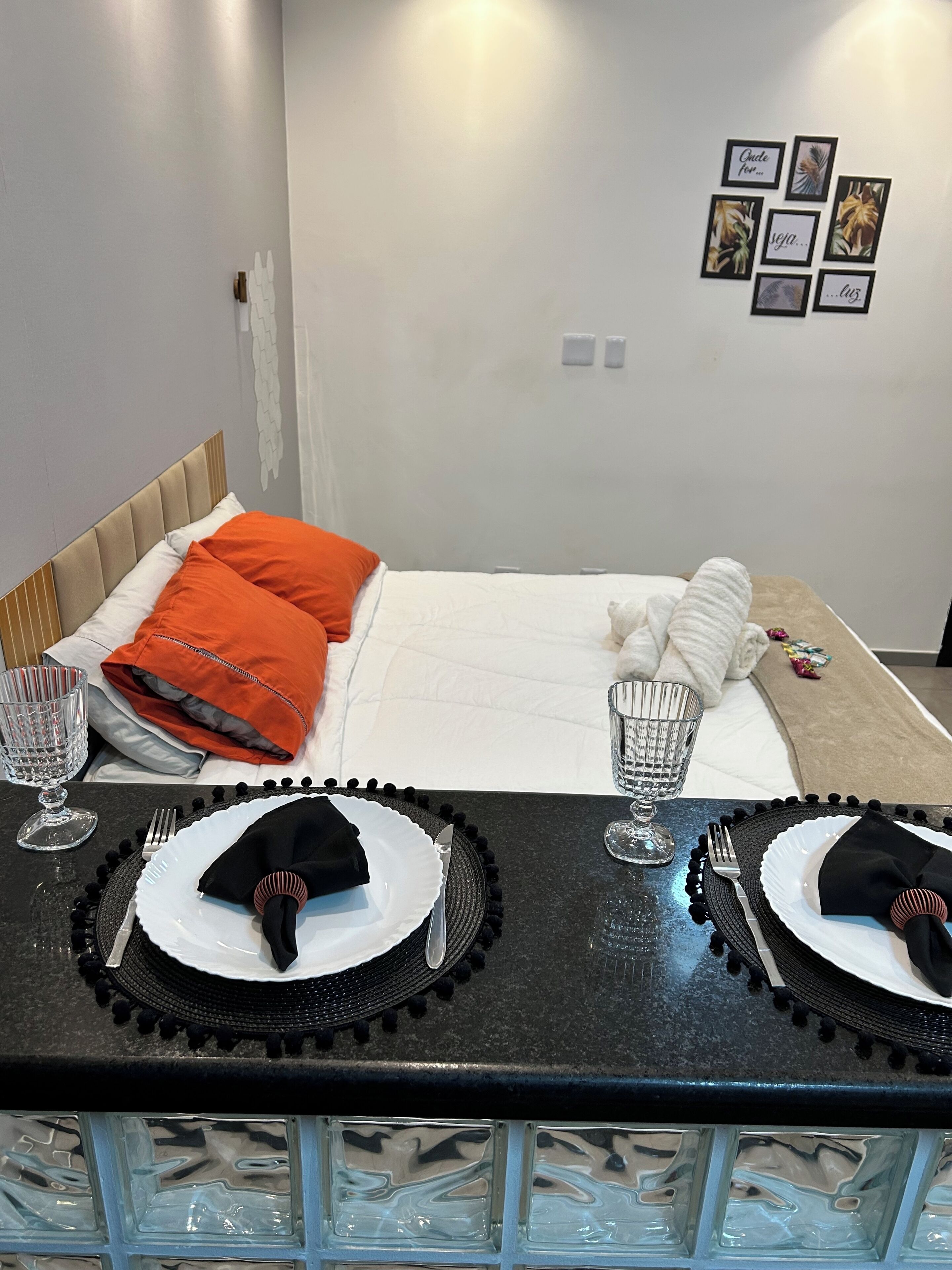 A neatly made bed with orange and white pillows, a bedside table with a glass and a metal tray, and a wall with framed pictures.
