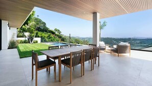 Eine moderne Außenpatio mit Holztisch und -stühlen, einer Glasreling und Blick auf einen Pool und Grünflächen.