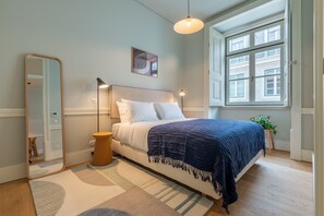 Een slaapkamer met een bed, nachtkastje, lamp, spiegel, raam en een plant.