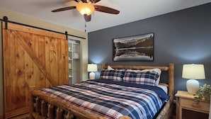 A bedroom with a wooden bed frame, a ceiling fan, a mounted painting, and a nightstand with a lamp. - Charming 3-bedroom cabin in Woodruff perfect for relaxing getaways (Woodruff)