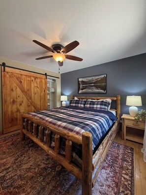 A bedroom with a wooden bed frame, a ceiling fan, a mounted painting, and a nightstand with a lamp.