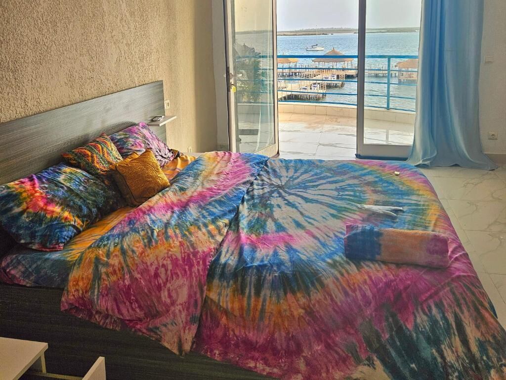 Una cama con ropa de cama colorida y vista a un puerto deportivo a través de un ventanal.