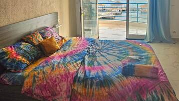 Una cama con ropa de cama colorida y vista a un puerto deportivo a través de un ventanal.