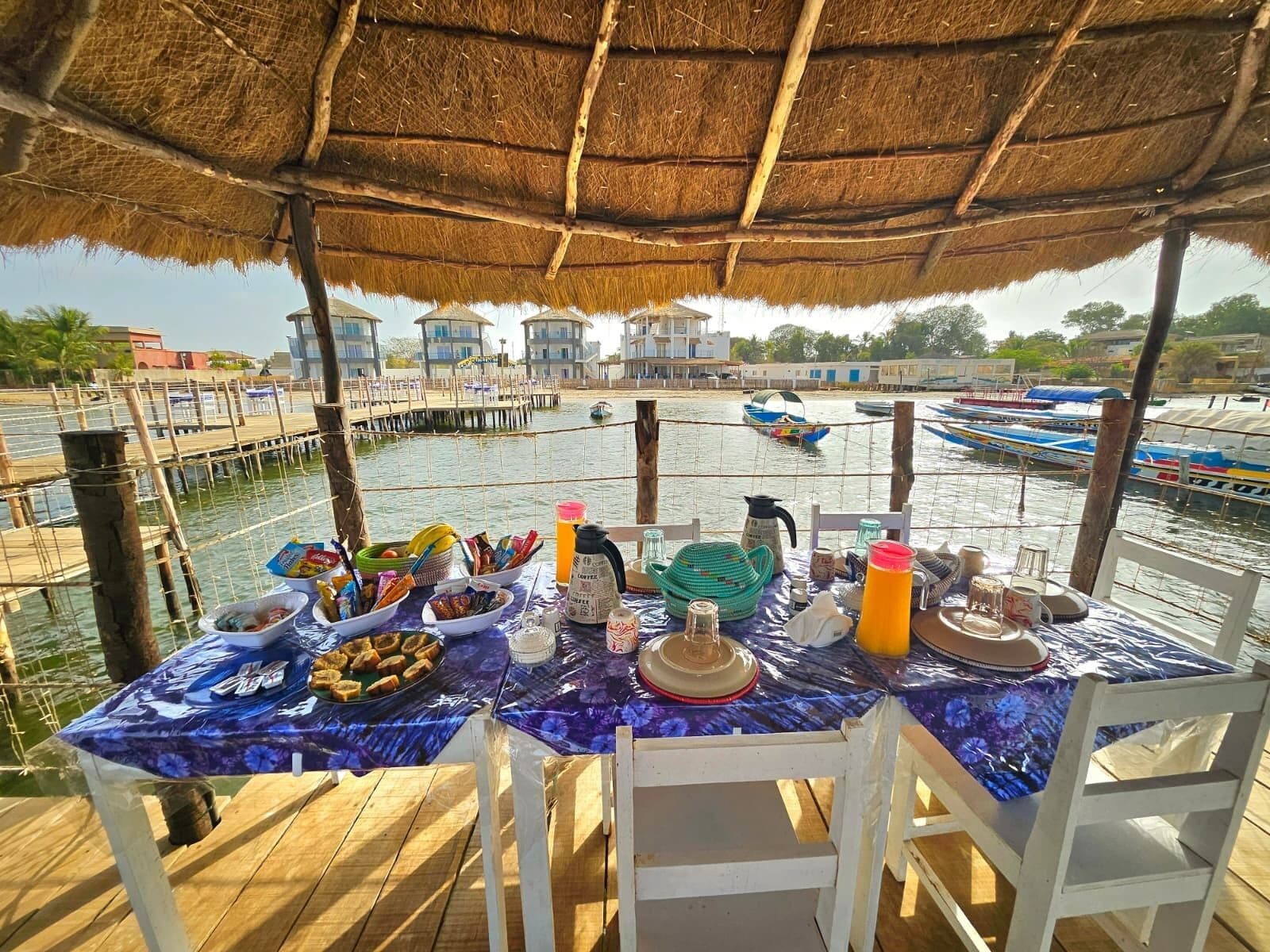 A table set up on a wooden deck with a thatched roof, overlooking a water body with boats and buildings in the background.