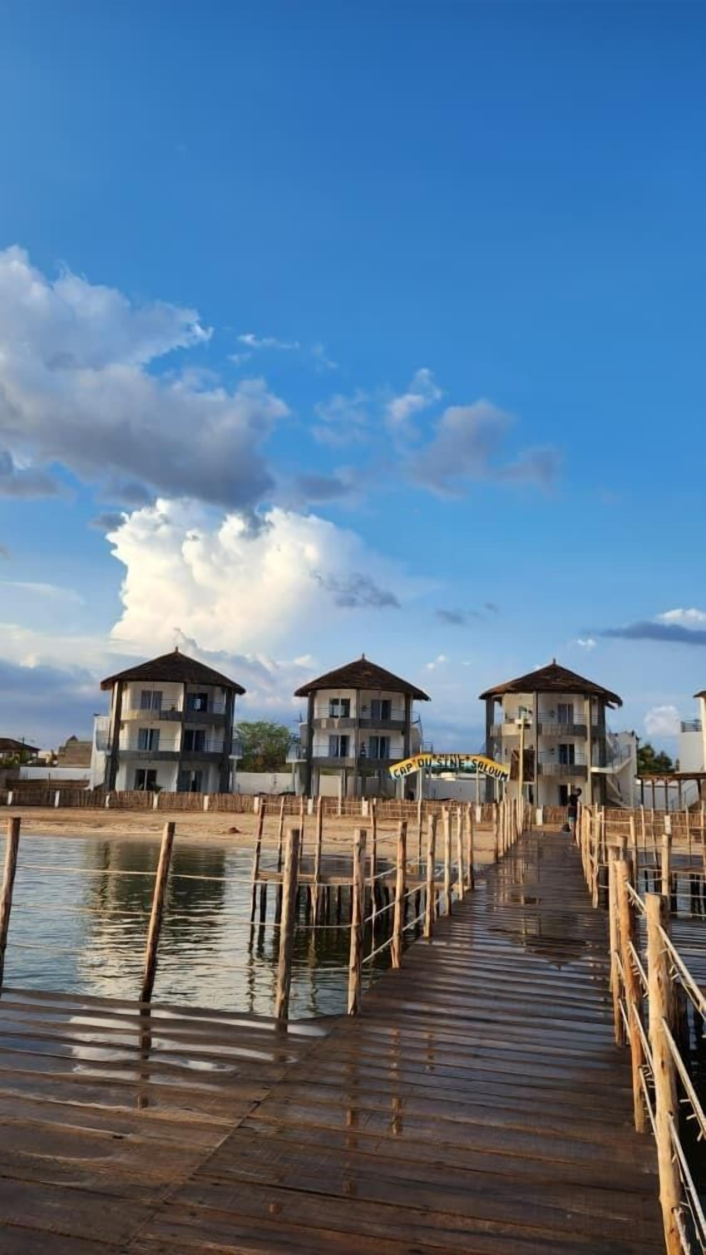 Un muelle de madera que lleva a una hilera de edificios con balcones, bajo un cielo parcialmente nublado.