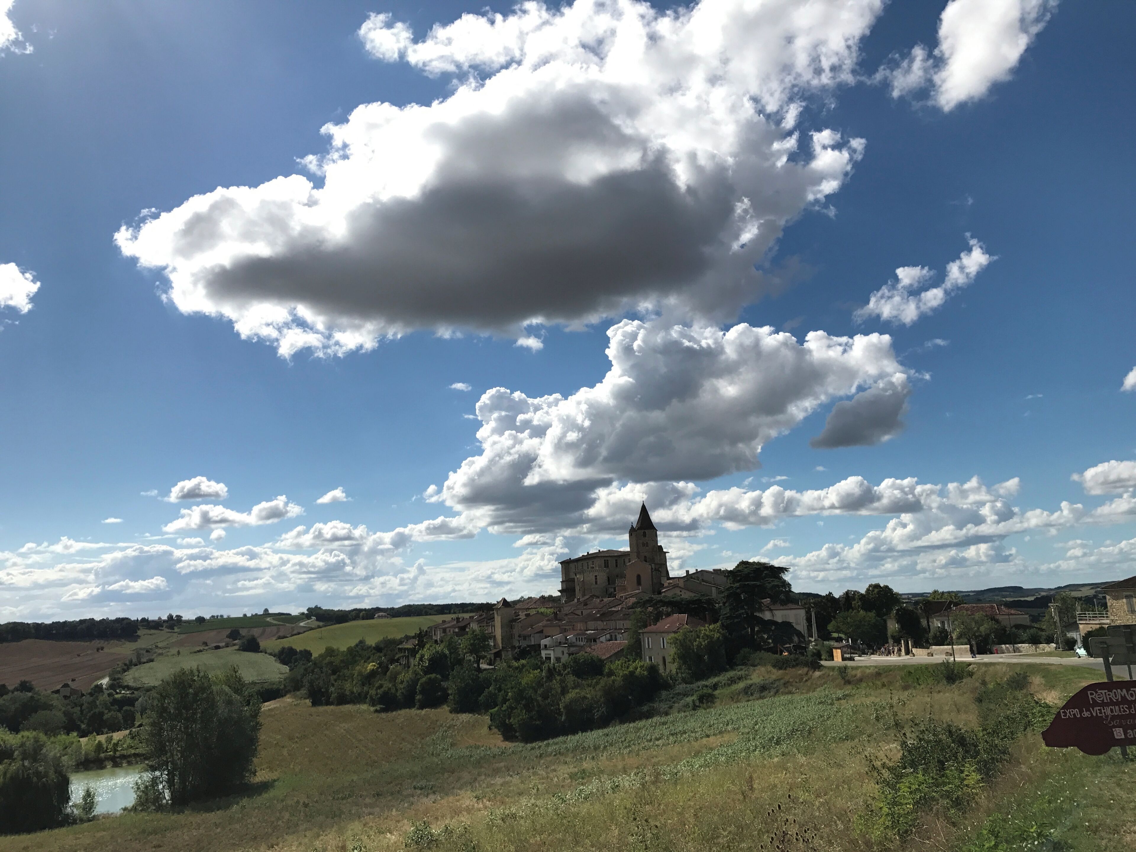 Un edificio storico con una torre, circondato da vegetazione e campi, sotto un cielo parzialmente nuvoloso.