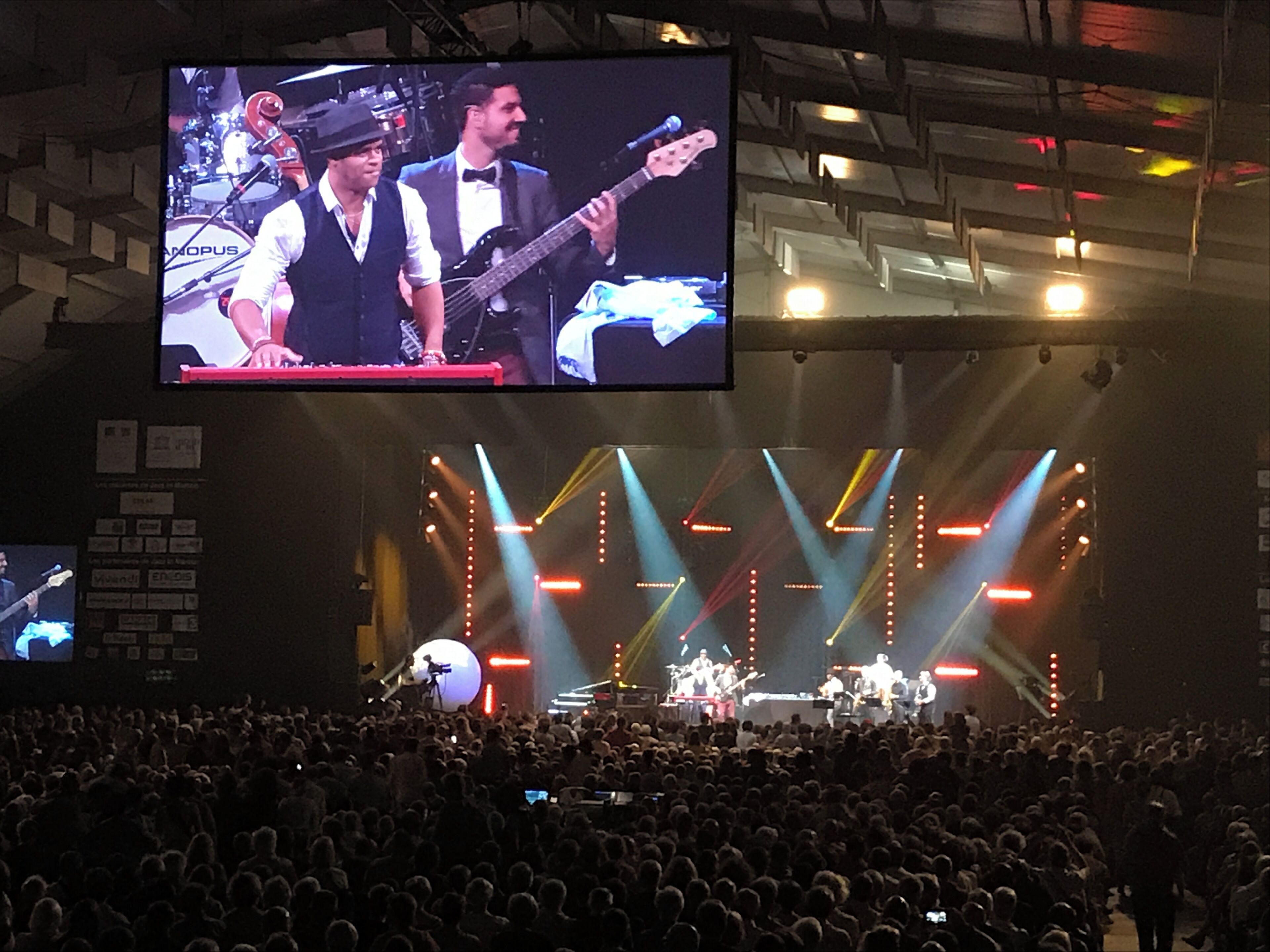 A concert stage with a band, a large screen above, and a crowd of people.