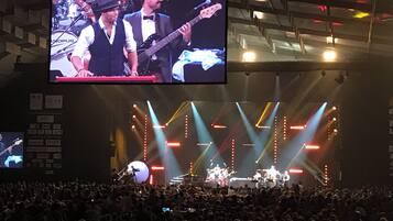 A concert stage with a band, a large screen above, and a crowd of people.