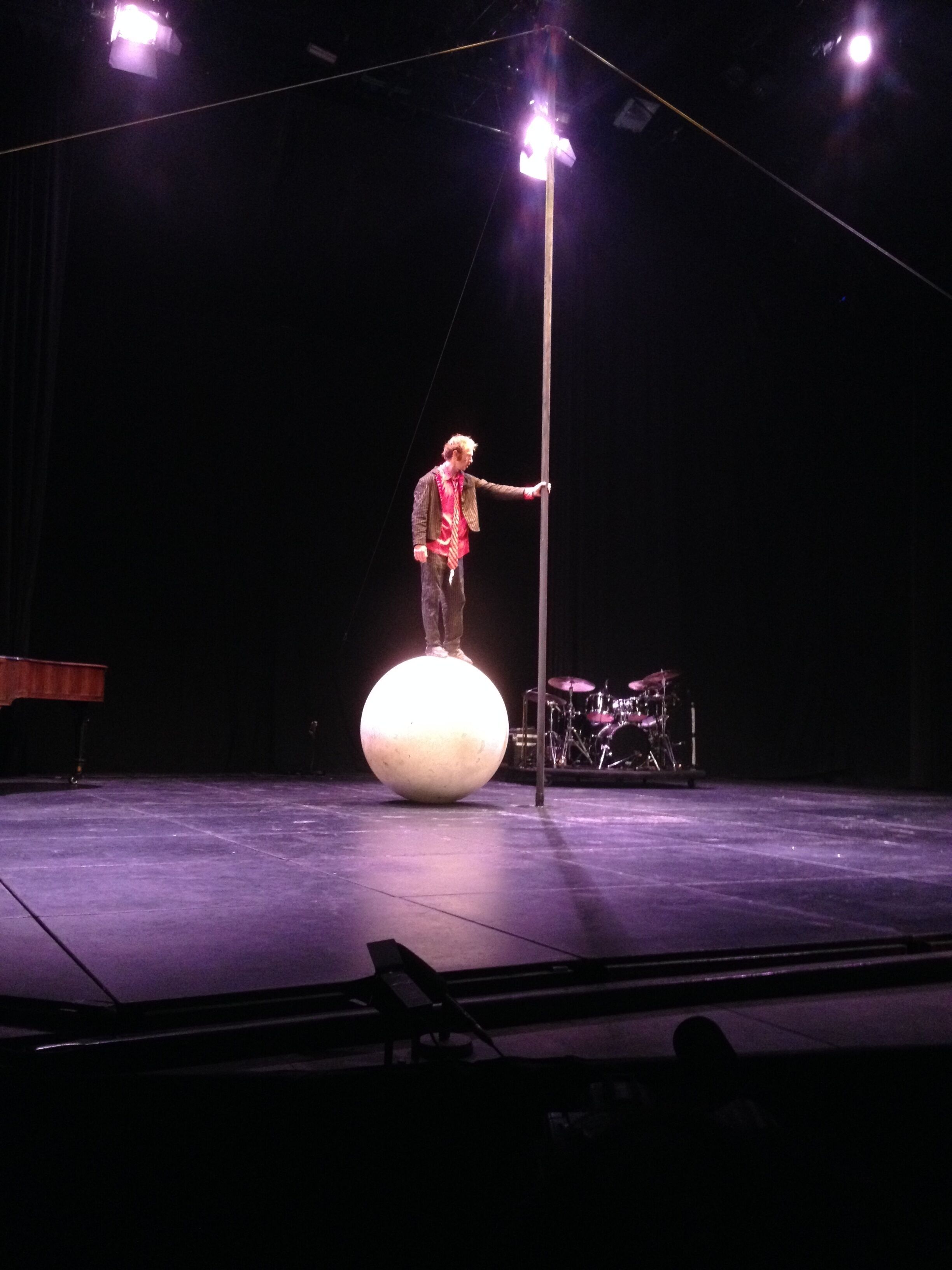 A stage with a performer balancing on a large sphere, a drum set, and a piano in the background.