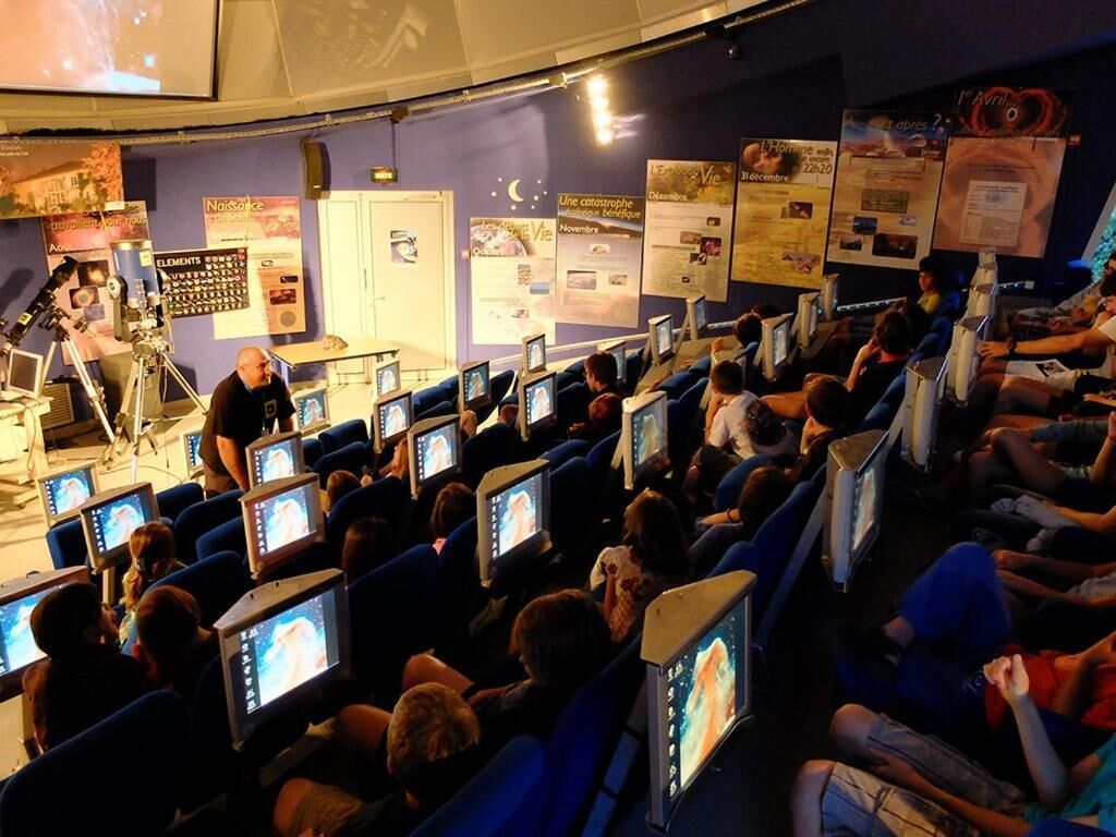 A lecture hall with rows of seats, a stage with a speaker, and educational posters on the walls.