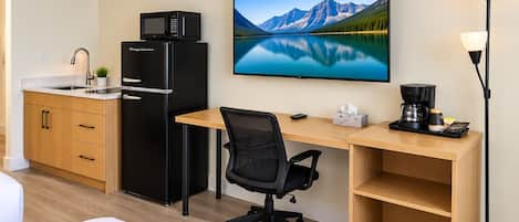 Habitación de hotel con kitchenette, escritorio con computadora, silla, cama y una ventana grande con vista a las montañas.