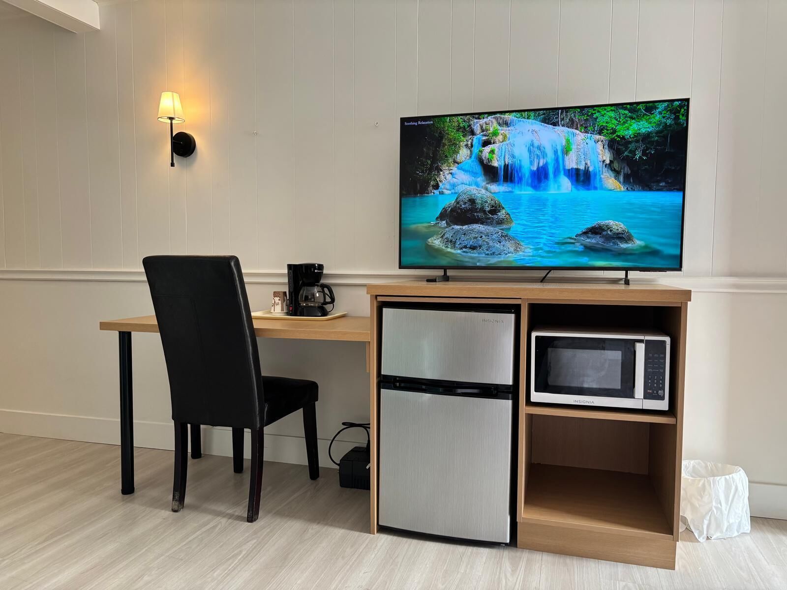 Une chambre moderne équipée d’un bureau en bois, d’une chaise noire, d’un téléviseur diffusant une scène de cascade, d’une machine à café, d’un mini-réfrigérateur et d’un four à micro-ondes.
