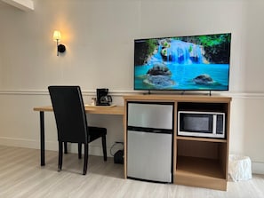 Une chambre moderne équipée d’un bureau en bois, d’une chaise noire, d’un téléviseur diffusant une scène de cascade, d’une machine à café, d’un mini-réfrigérateur et d’un four à micro-ondes.