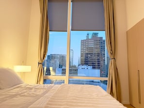 A hotel room with a bed, a window with curtains, and a view of city buildings.