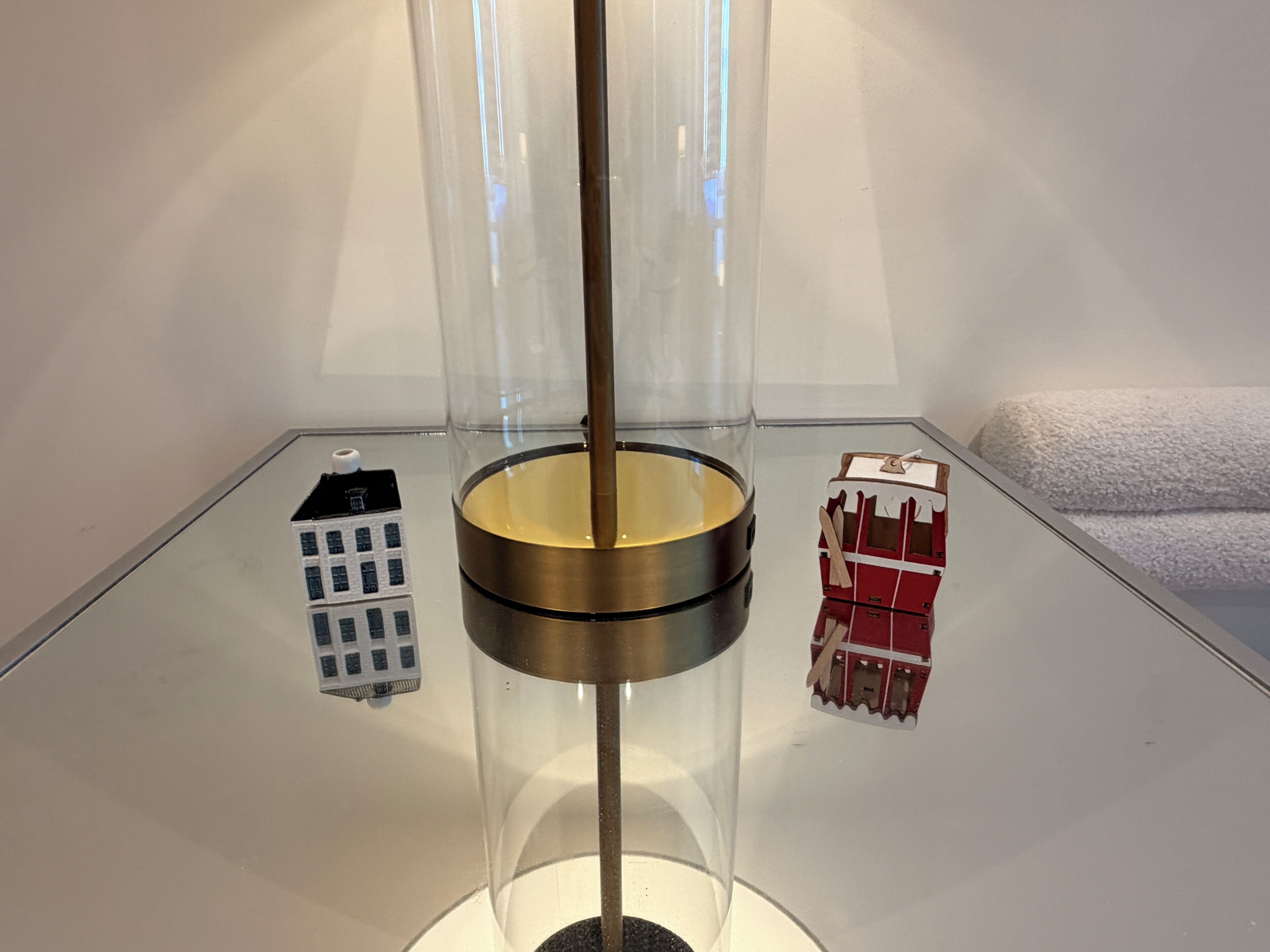 A transparent cylindrical object with a metallic base, a small building model, and a wrapped item on a reflective surface.