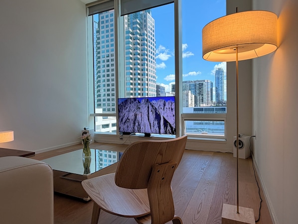 A modern living room with a large window, a flat-screen TV, a wooden chair, a floor lamp, and a vase with flowers.
