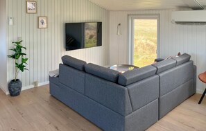 A modern living room with a grey sectional sofa, a flat-screen TV, a potted plant, and a view of the outdoors through a sliding door. - Beautiful home in Lemvig with sauna (Lemvig)