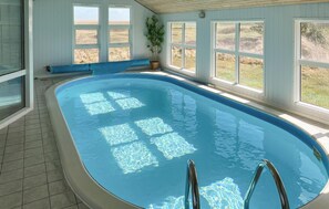 An indoor swimming pool with clear blue water, a wooden deck, and large windows offering views of the outdoors. - Beautiful home in Lemvig with sauna (Lemvig)