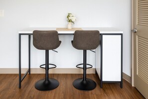 Et moderne værelse med et hvidt skrivebord, to grå barstole, en lille vase med hvide blomster og trægulv.