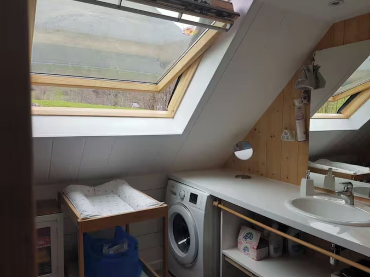 Una habitación compacta, con techo inclinado, tragaluz, lavadora, lavabo y espejo.