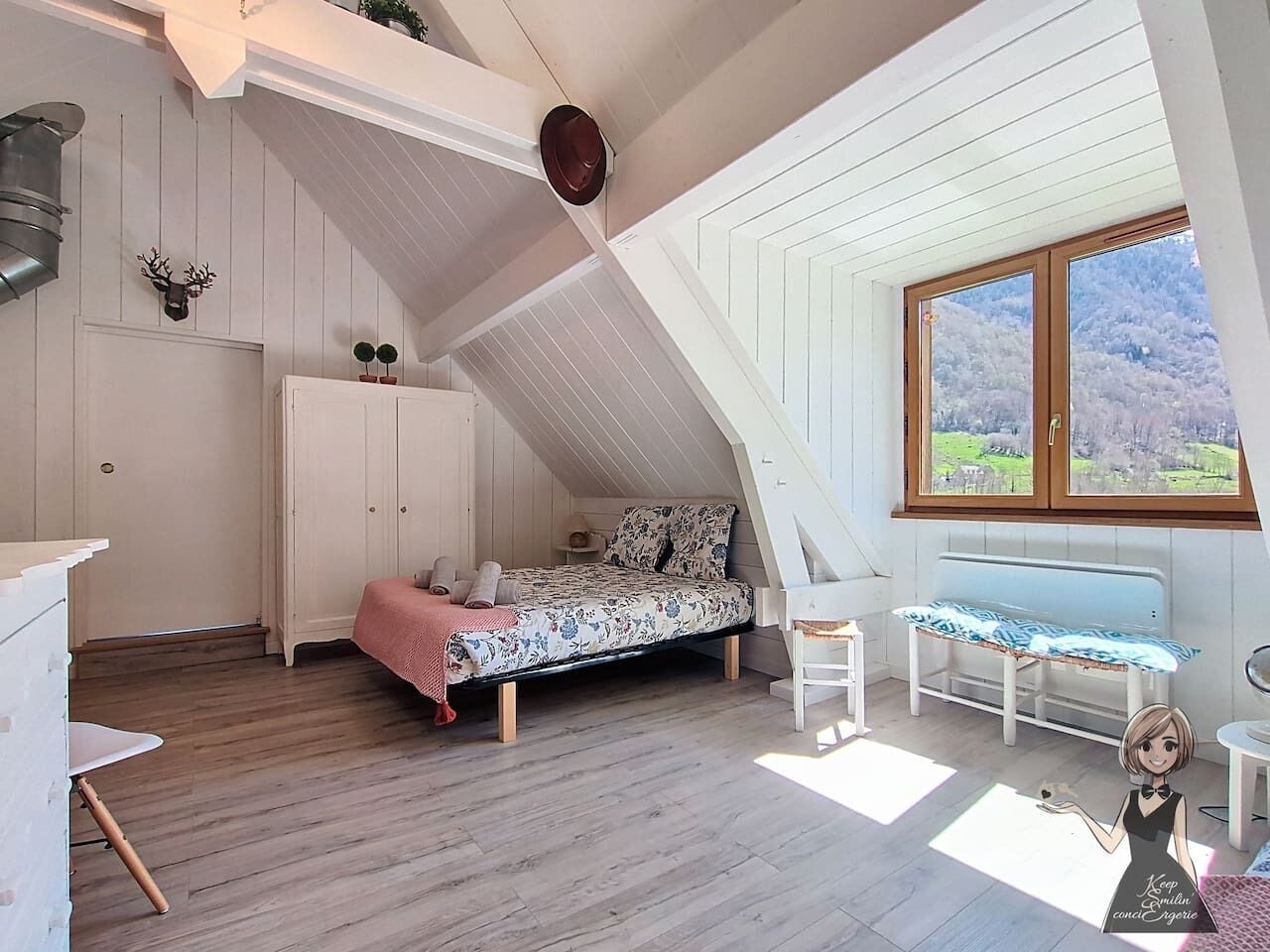 Een slaapkamer met een schuine kap, houten vloeren, een bed met bloemenprint beddengoed, een bankje, een klein tafeltje en een raam met uitzicht op groen.