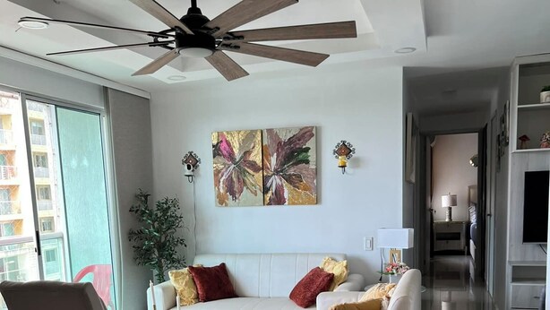 A modern living room with a ceiling fan, a white sofa with red and gold cushions, a glass coffee table, and a large abstract painting on the wall. - Spacious 3-bedroom apartment in perfect Barranquilla with AC (Barranquilla)