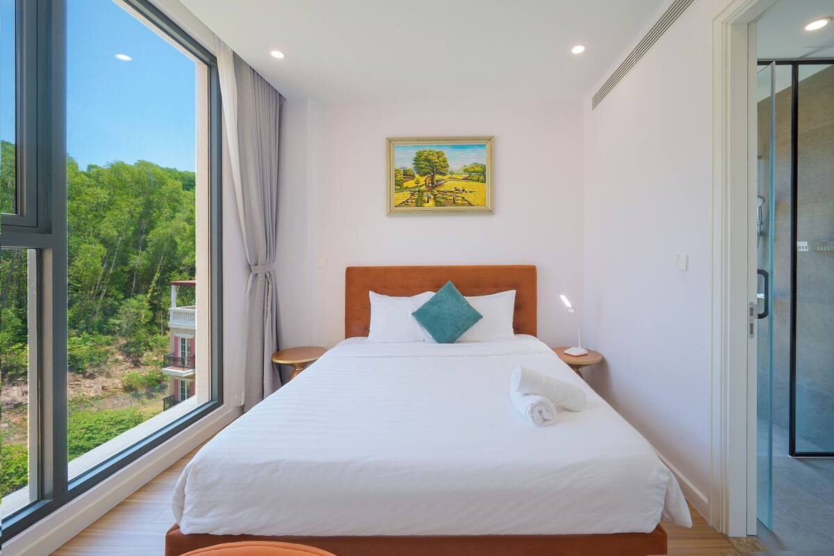 Habitación de hotel con un ventanal grande, una cama con ropa blanca, un cuadro en la pared y una puerta de cristal que da a la zona de la ducha.