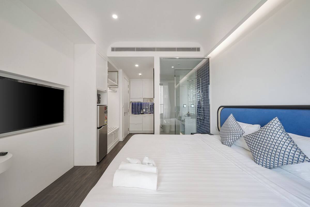 Una habitación de hotel moderna con una cama grande, un televisor de pantalla plana en la pared, un vestidor y un baño con cabina de ducha de vidrio.