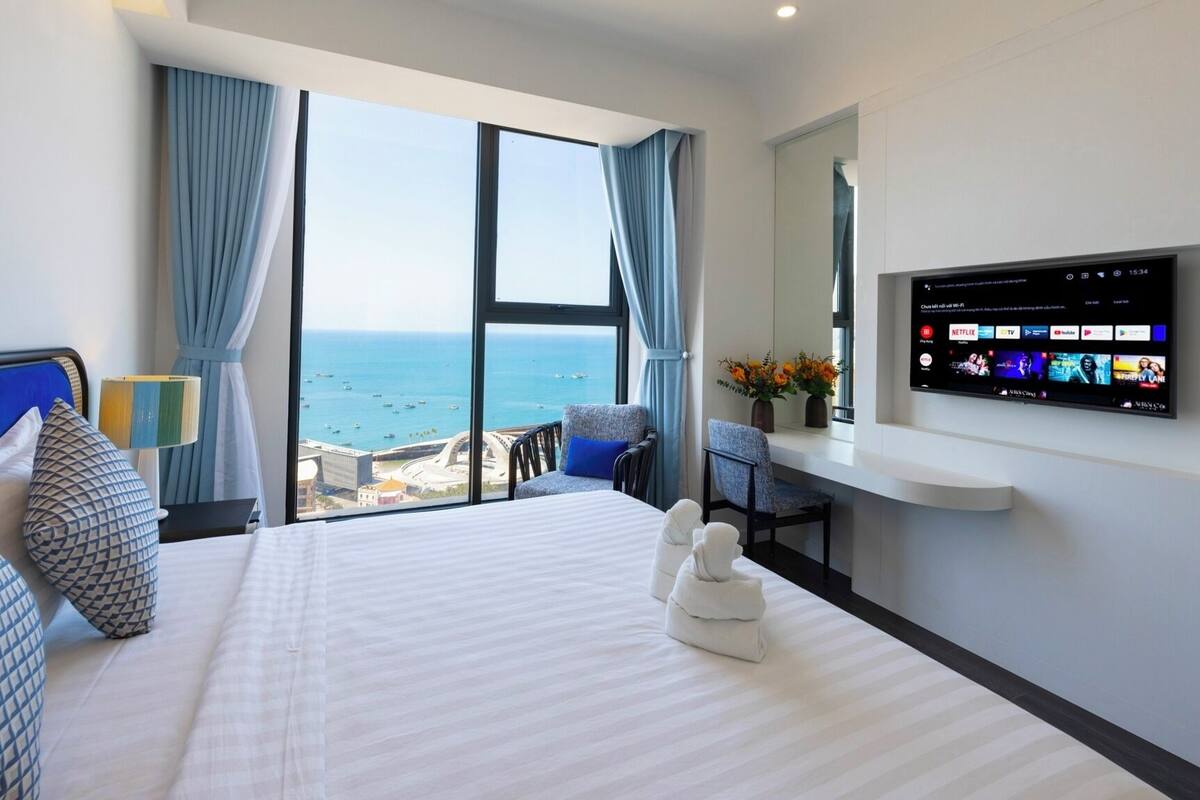 Habitación de hotel con una cama grande, un televisor montado en la pared, vista al mar y un balcón con una silla.