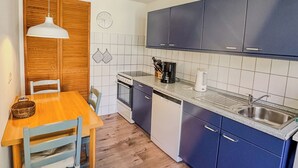 Et kompakt køkken med træskabe, en indbygget ovn, en opvaskemaskine og et spiseområde med et træbord og stole.